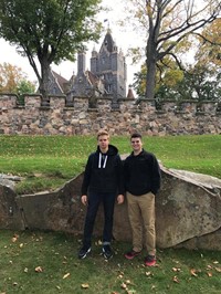 two male students smile standing by rock with large old fashioned building in background