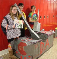 three students standing with cardboard boat